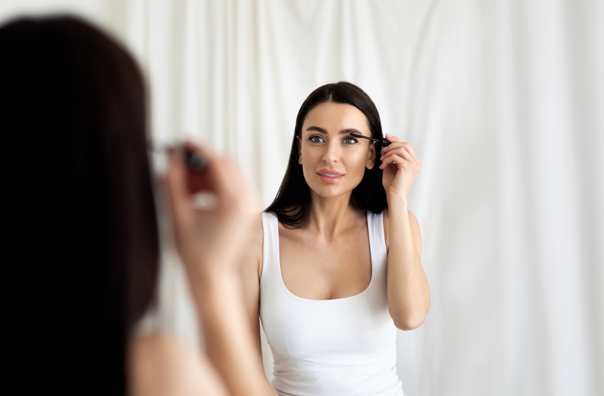 Smiling millennial cute pretty lady with healthy shining skin puts mascara on eyelashes